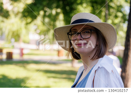 Young woman enjoys sunny day in park, wearing stylish hat and glasses, surrounded by greenery 130410257