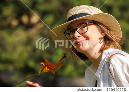 Woman wearing a sun hat smiles while holding a flower in a lush green park during sunny weather Woman wearing a sun hat smiles while holding a flower in a lush green park during sunny weather 130410258