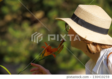 Woman enjoying a warm day in nature while holding a vibrant flower near a blooming garden Woman enjoying a warm day in nature while holding a vibrant flower near a blooming garden 130410259