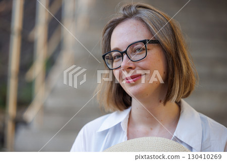 Young woman with glasses smiling while holding a straw hat on outdoor stairs Young woman with glasses smiling while holding a straw hat on outdoor stairs 130410269