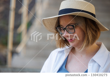 Woman in a straw hat enjoying a quiet moment outdoors near stairs on a sunny day 130410273