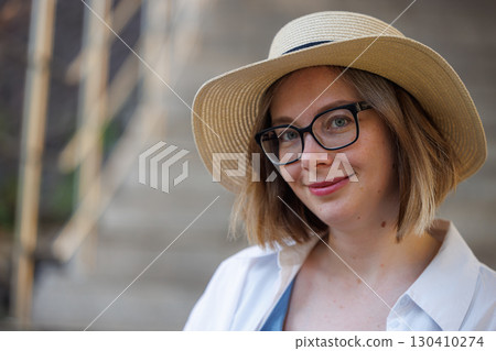 Woman with short hair and glasses wearing a hat smiles on outdoor stairs in soft lighting during the day Woman with short hair and glasses wearing a hat smiles on outdoor stairs in soft lighting during the day 130410274