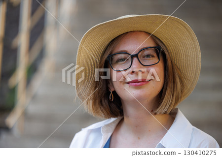 Smiling woman in glasses and sunhat poses on outdoor stairs in summer afternoon light 130410275