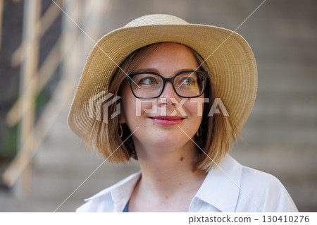 Woman wearing a hat and glasses smiling while standing near a staircase in a sunny outdoor setting 130410276