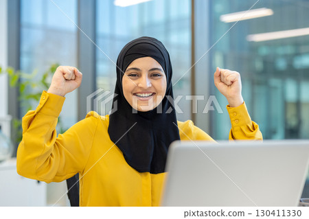 Happy businesswoman wearing a hijab raising her arms in celebration, surrounded by a modern office environment, rejoicing over a successful project and recent achievement in front of her laptop 130411330