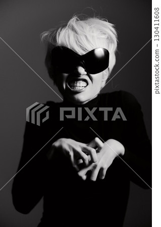 Confident young woman with short blonde hair wearing oversized sunglasses and black outfit, expressing fierce emotion against a dark background. Fashion and attitude concept. 130411608