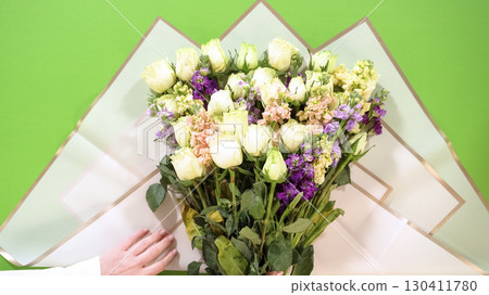 Two hands adjust a bouquet position over wrapping paper before folding. The image is captured overhead on a green surface for contrast. Two hands adjust a bouquet position over wrapping paper before folding. The image is captured overhead on a green surface for contrast. 130411780