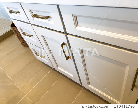 Lower bathroom cabinets with brass hardware sit on tan tile flooring. The design features shaker-style doors and a clean modern finish. Lower bathroom cabinets with brass hardware sit on tan tile flooring. The design features shaker-style doors and a clean modern finish. 130411786