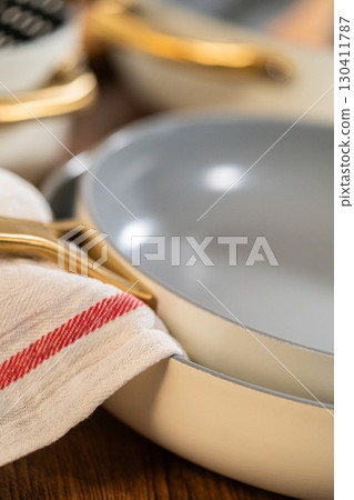 Two non-stick pans stacked together with a tea towel draped over the edge. The setup is arranged on a wooden table with warm lighting. Two non-stick pans stacked together with a tea towel draped over the edge. The setup is arranged on a wooden table with warm lighting. 130411787
