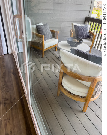 Cushioned patio chairs and a table are visible through a glass sliding door, with a dark composite deck underfoot. The perspective offers an inside-out view of a cozy backyard setup. 130411788