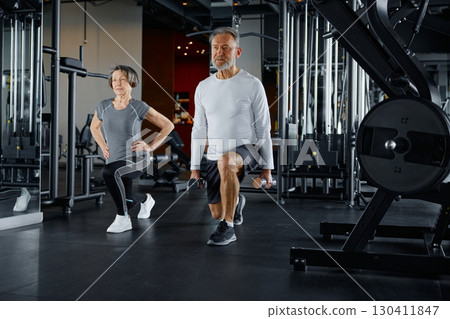 Elderly man and a woman in athletic attire doing lunges in a gym 130411847