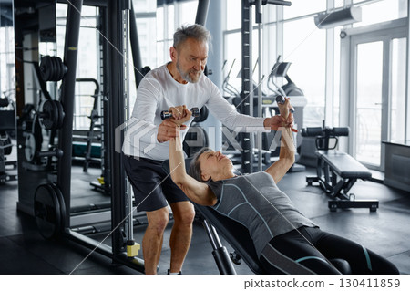Mature man helps a mature woman lift dumbbells on weight bench in a gym 130411859