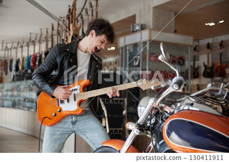 Expressive man in a leather jacket is playing guitar in a store Expressive man in a leather jacket is playing guitar in a store 130411911