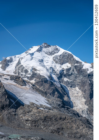 Piz Bernina stands prominently in the Swiss Alps, showcasing its snow-covered peak against a bright blue sky. Glacial formations surround this iconic mountain in the Bernina range. Piz Bernina stands prominently in the Swiss Alps, showcasing its snow-covered peak against a bright blue sky. Glacial formations surround this iconic mountain in the Bernina range. 130412609