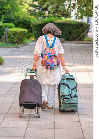 Senior woman walking away on urban sidewalk with two travel bags and colorful backpack. Concept of independent travel, solo journey, senior adventure 130414430