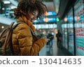 Smiling young woman in an airport terminal looking at her phone, with a backpack on her shoulder and a flight information display board in the blurry background 130414635