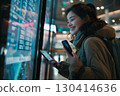 Smiling young woman in an airport terminal looking at her phone, with a backpack on her shoulder and a flight information display board in the blurry background 130414636