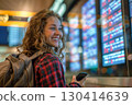 Smiling young woman in an airport terminal looking at her phone, with a backpack on her shoulder and a flight information display board in the blurry background 130414639