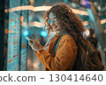 Smiling young woman in an airport terminal looking at her phone, with a backpack on her shoulder and a flight information display board in the blurry background 130414640