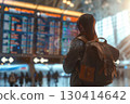 Smiling young woman in an airport terminal looking at her phone, with a backpack on her shoulder and a flight information display board in the blurry background 130414642