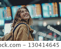 Smiling young woman in an airport terminal looking at her phone, with a backpack on her shoulder and a flight information display board in the blurry background 130414645