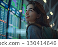 Smiling young woman in an airport terminal looking at her phone, with a backpack on her shoulder and a flight information display board in the blurry background 130414646