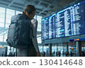 Smiling young woman in an airport terminal looking at her phone, with a backpack on her shoulder and a flight information display board in the blurry background 130414648