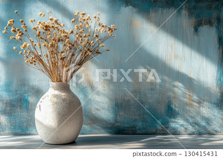 White ceramic vase with dried golden flowers on a wooden table, against a textured blue wall with natural sunlight and shadow patterns. 130415431