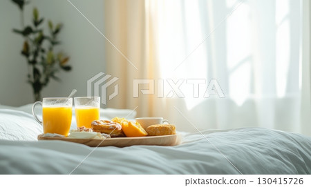 Two mugs of orange juice, pastries, orange slices, and coffee cup served on wooden tray placed on comfortable bed with white sheets in sunlit hotel room creating cozy and inviting atmosphere 130415726