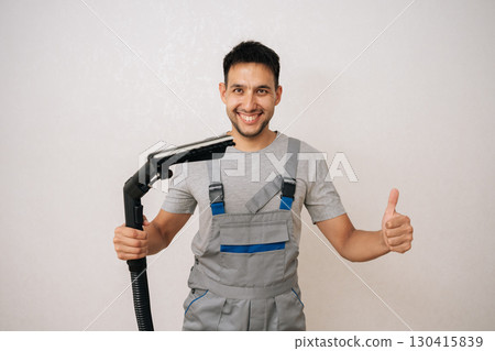 Portrait of cheerful professional cleaning service employee in overall holding washing vacuum cleaner, getting ready to cleaning in living room, smiling looking at camera, showing thumbs up gesture. 130415839