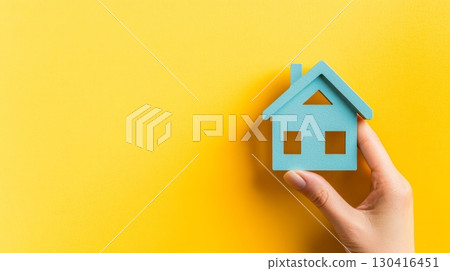 Hand holds a small blue house against a vibrant yellow backdrop, representing real estate investment, homeownership, and the mortgage process Hand holds a small blue house against a vibrant yellow backdrop, representing real estate investment, homeownership, and the mortgage process 130416451