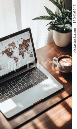Open laptop displaying world map on wooden desk next to cappuccino and potted plant, suggesting online travel planning or global business strategies in a cozy work environment 130416943