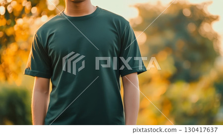Close up of a man wearing a plain green t shirt, providing a clean template for apparel design or branding, with a blurred outdoor background featuring warm sunset light filtering through trees Close up of a man wearing a plain green t shirt, providing a clean template for apparel design or branding, with a blurred outdoor background featuring warm sunset light filtering through trees 130417637