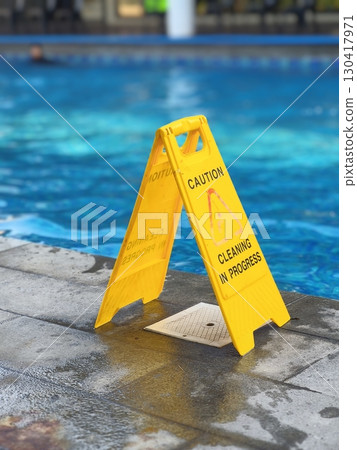 Yellow caution sign on wet poolside surface near swimming pool, warning of cleaning in progress and slippery floor hazard for safety awareness. Yellow caution sign on wet poolside surface near swimming pool, warning of cleaning in progress and slippery floor hazard for safety awareness. 130417971