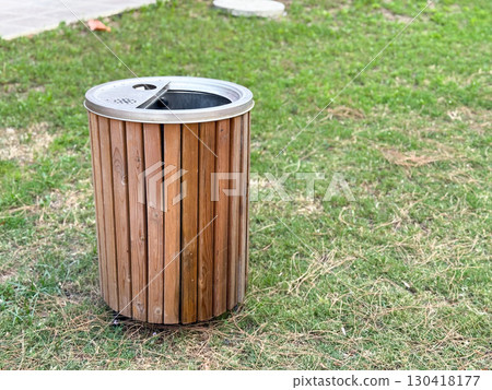 Wooden trash bin with metal lid stands on grass in public park, copy space. Ecology, sustainability, and waste management reflecting cleanliness, urban service and environmental care outdoors. Wooden trash bin with metal lid stands on grass in public park, copy space. Ecology, sustainability, and waste management reflecting cleanliness, urban service and environmental care outdoors. 130418177