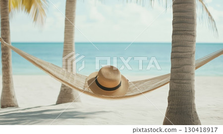 Straw hat lies on a hammock strung between two palm trees on a white sand beach, overlooking a tranquil turquoise ocean, evoking a sense of peaceful relaxation and tropical getaway 130418917