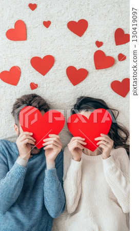 Young couple lying on carpet hiding their faces with red paper hearts surrounded by other small hearts celebrating valentine's day, love, and spring Young couple lying on carpet hiding their faces with red paper hearts surrounded by other small hearts celebrating valentine's day, love, and spring 130419097