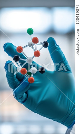 Hand wearing a blue glove holding a molecular model made of transparent sticks and colored balls, representing a chemical compound or molecule, in a scientific laboratory setting 130419872