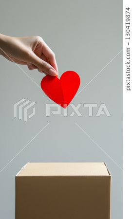 Hand holding a red paper heart, about to drop it into a cardboard donation box, symbolizing charity, healthcare donations, or organ donation, against a light gray background 130419874