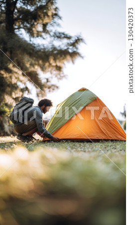 Backpacker setting up an orange and green tent on lush grass in a tranquil campground surrounded by trees, basking in the warm glow of a breathtaking sunset 130420373