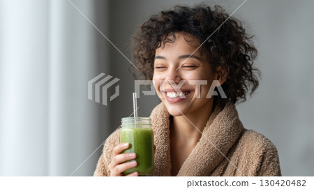 Happy young woman wearing bathrobe is holding a glass jar with reusable straw filled with green detox smoothie, enjoying healthy lifestyle and smiling with closed eyes 130420482