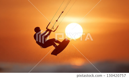 Silhouette of kitesurfer performing acrobatic tricks in the air against a vibrant orange sunset over the sea, showcasing the thrill and beauty of watersports 130420757