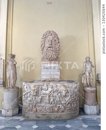 VATICAN, ROME, ITALY - AUGUST 02, 2025 Marble head of a bearded deity possibly Jupiter or Saturn. Mythological figure displayed in the Chiaramonti Museum stands on a sarcophagus with Dionysian scene VATICAN, ROME, ITALY - AUGUST 02, 2025 Marble head of a bearded deity possibly Jupiter or Saturn. Mythological figure displayed in the Chiaramonti Museum stands on a sarcophagus with Dionysian scene 130420844