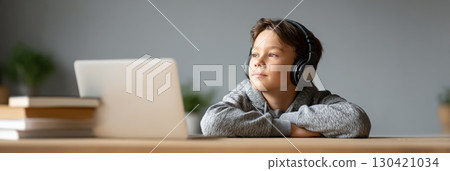 Schoolboy wearing headphones is attending online lesson using laptop and looking away while sitting at desk at home with books nearby, enjoying remote learning and homeschooling 130421034