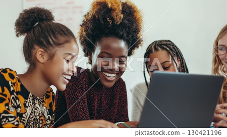 Four cheerful female students collaborating on a school project, using a laptop and smiling while enjoying the learning process together in a classroom 130421196