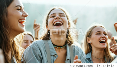 Group of young women laughing joyfully at an outdoor event, showcasing happiness and camaraderie, surrounded by friends enjoying a lively atmosphere with sunlight filtering through 130421612