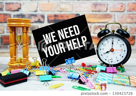 We Need Your Help text on paper card with alarm clock and stationary on wooden background 130423726