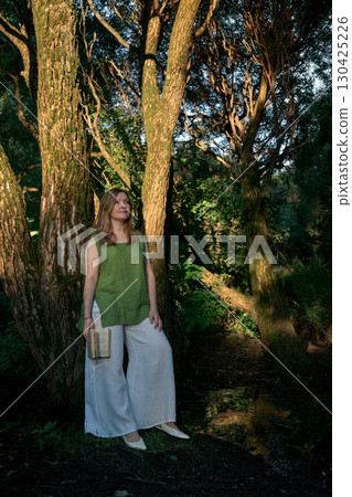 Woman in a green top and white trousers stands with a book in hand, leaning against a tree in Izmailovsky Park, Moscow Woman in a green top and white trousers stands with a book in hand, leaning against a tree in Izmailovsky Park, Moscow 130425226
