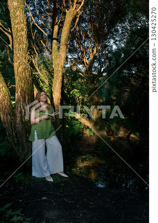 Woman in a green linen top and white trousers leans against a tree while holding an open book in Izmailovsky Park, Moscow Woman in a green linen top and white trousers leans against a tree while holding an open book in Izmailovsky Park, Moscow 130425270
