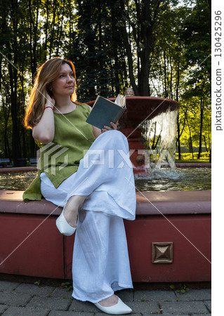 A young woman sits by a fountain in a park, holding an open book and gazing thoughtfully into the distance A young woman sits by a fountain in a park, holding an open book and gazing thoughtfully into the distance 130425296